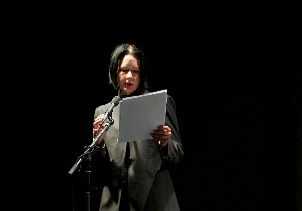 Vanessa Place reading from white papers in a black room