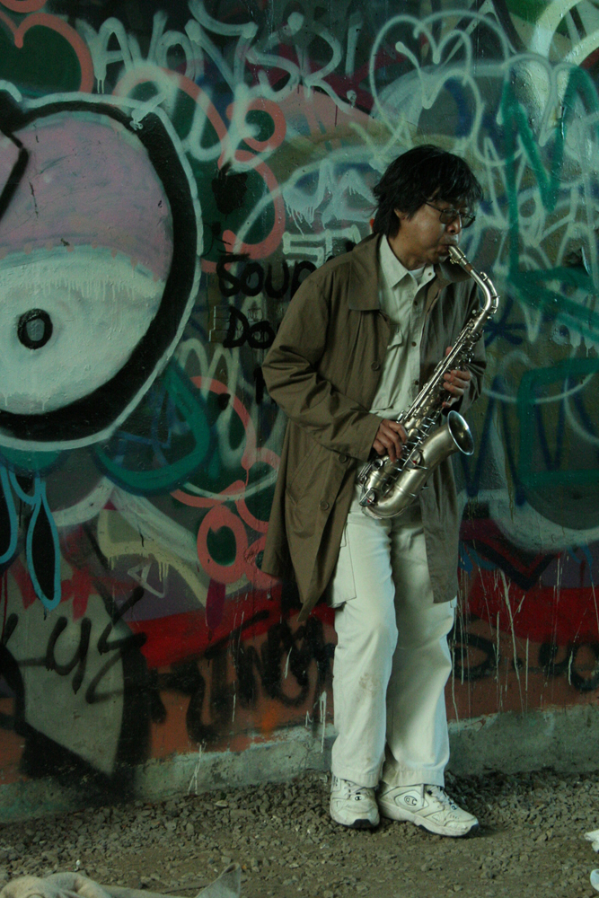 Tamio Shiraishi playing saxophone near graffiti covered walls, white jeans