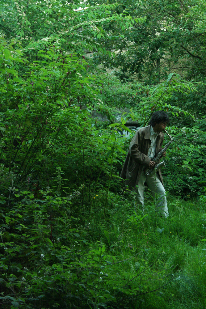 Tamio Shiraishi playing saxophone among leaves trees grasses and bushes