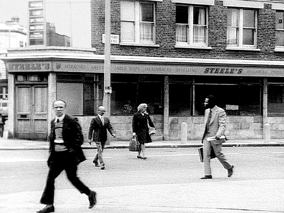 A B and W film still of several people crossing a street in 1960's london