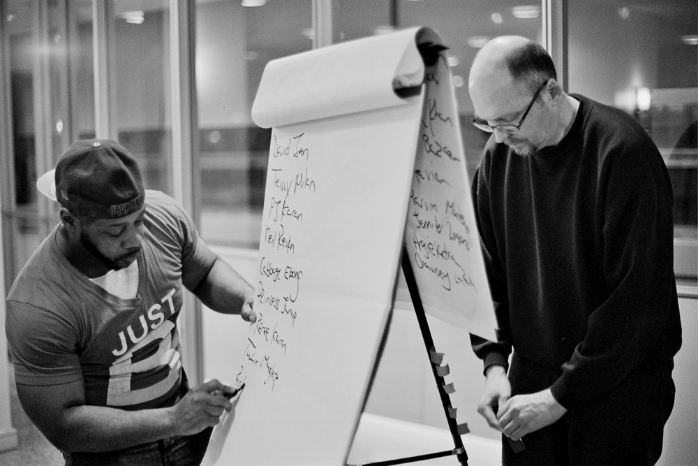 Two men stand at an flipchart stand as one in a cap writes on the paper