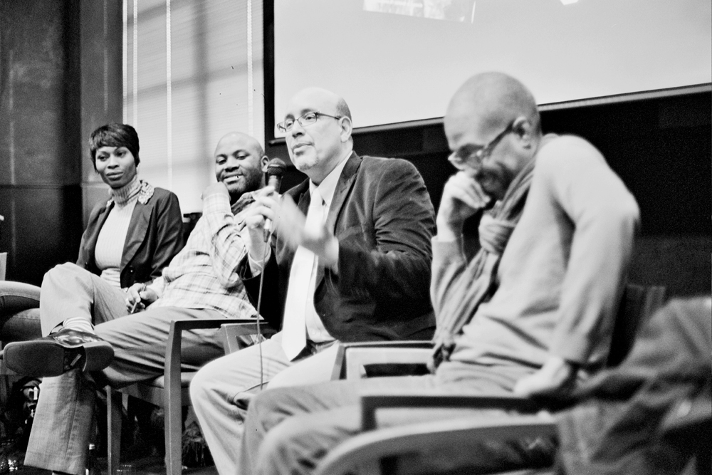 A panel of four folks sit as one wearing a dark jacket and specs speaks