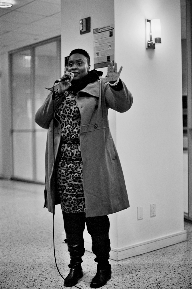 A woman in a mac and boots speaks on a mic whilst standing 