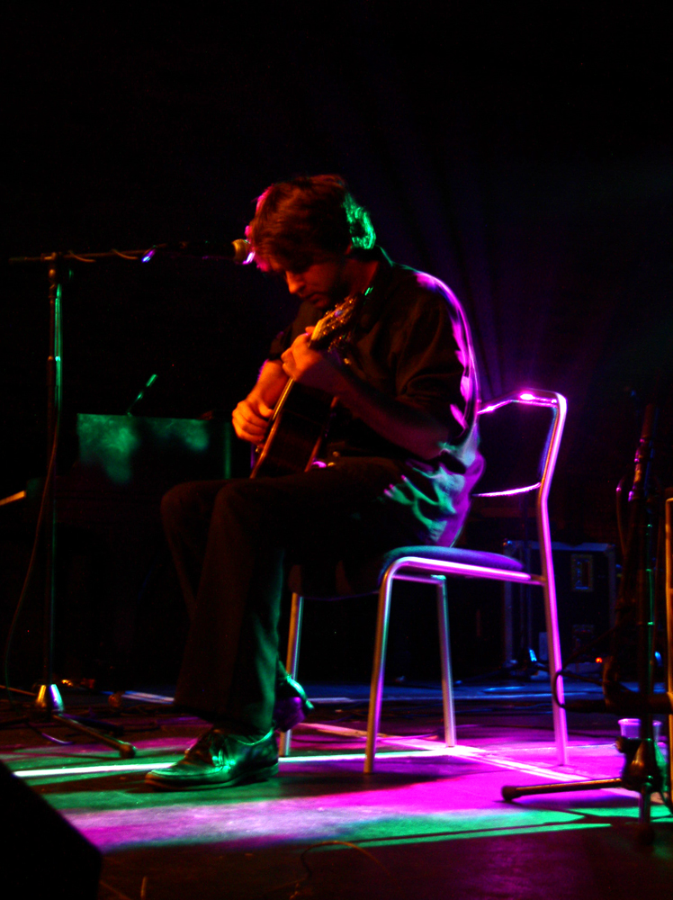 A seated Ben Chasney plays guitar on a sparsely illuminated stage