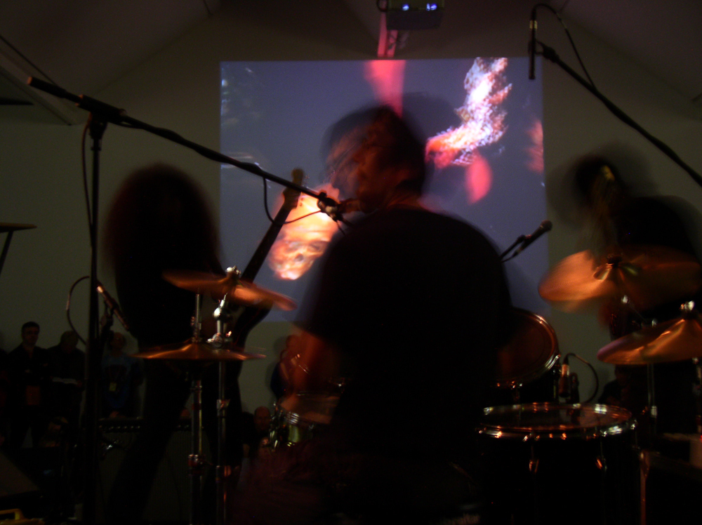 Three blurry dark band members play in front of a projection