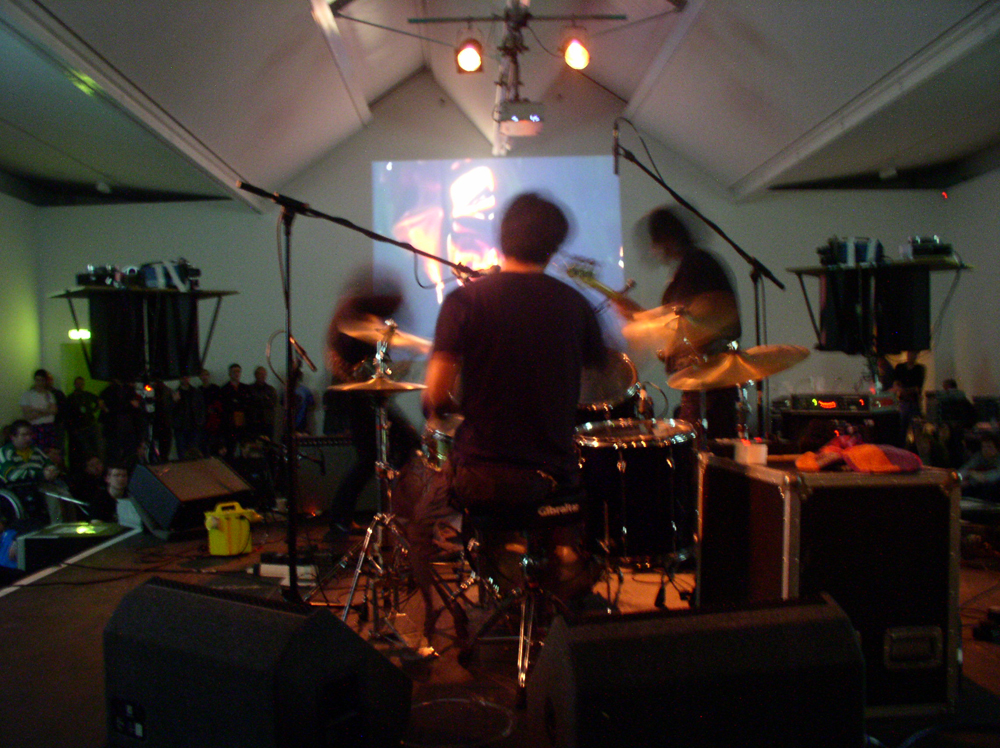 Three blurry dark band members play in front of a projection