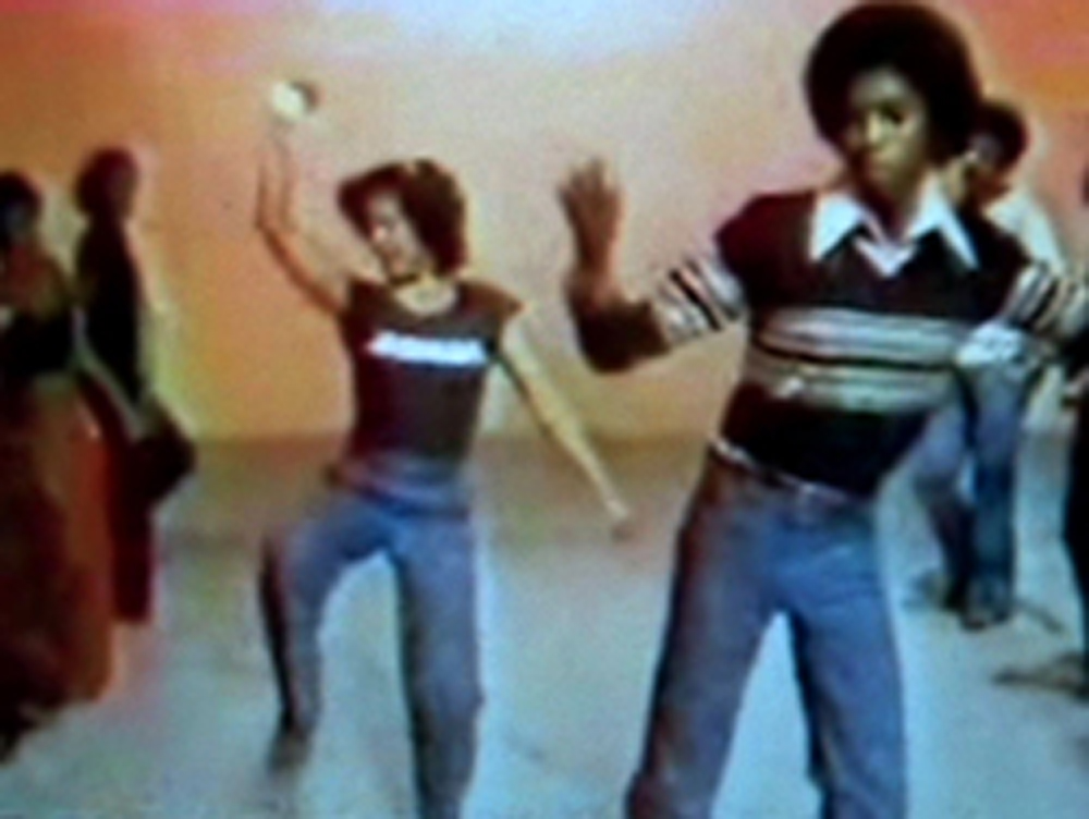 A video still of several folks pulling dance moves in front of an orange wall