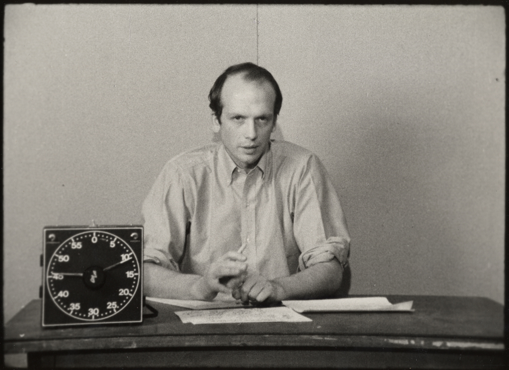 A man sits at a desk with a large timer clock on it and speaks to camera