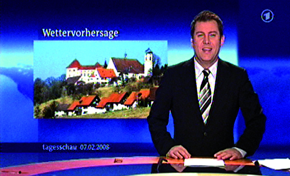 A newsreader in a suit looks to camera as a blue backdrop shows buildings