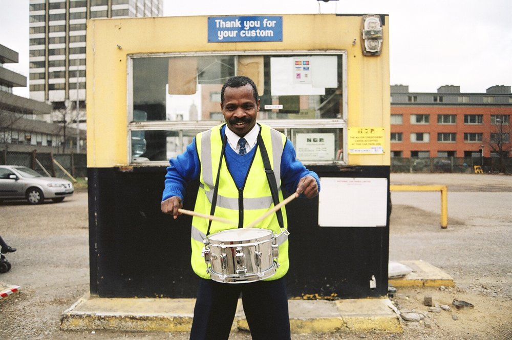 A man in Hi Vis smiles as he plays a snare drum in a car park