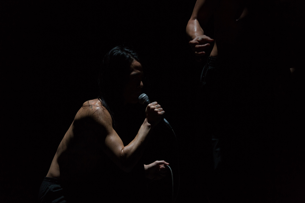 Crouched figure with microphone. They are in a dark room and their torso is lit