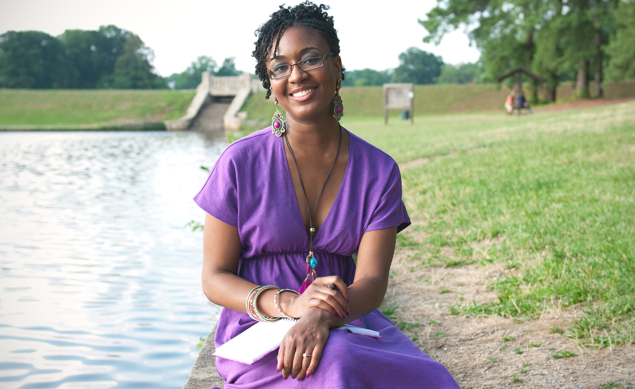 Camisha Jones smiles while sitting on a cement surface at the edge of a lake