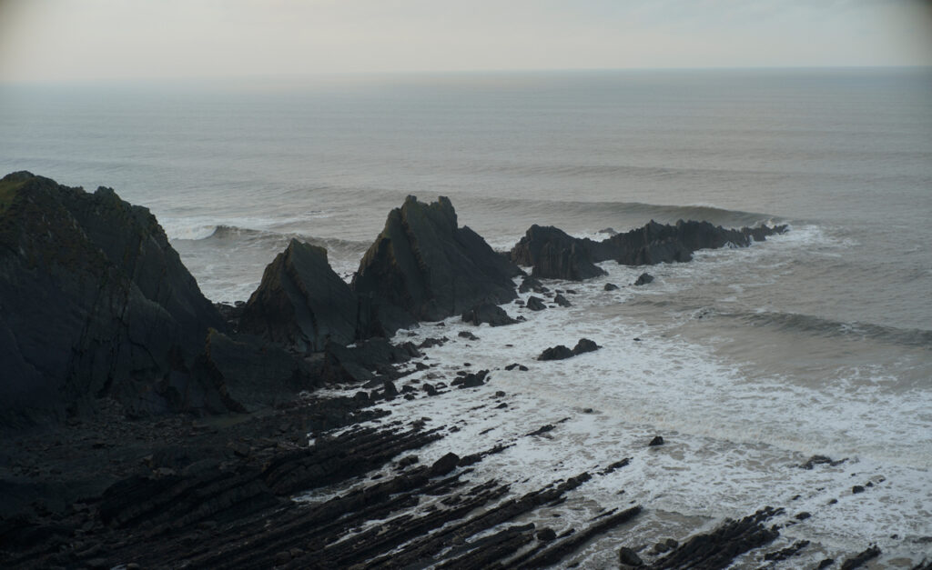 A jagged bluff of rocks juts out into a grey and moody sea as waves lap at shore