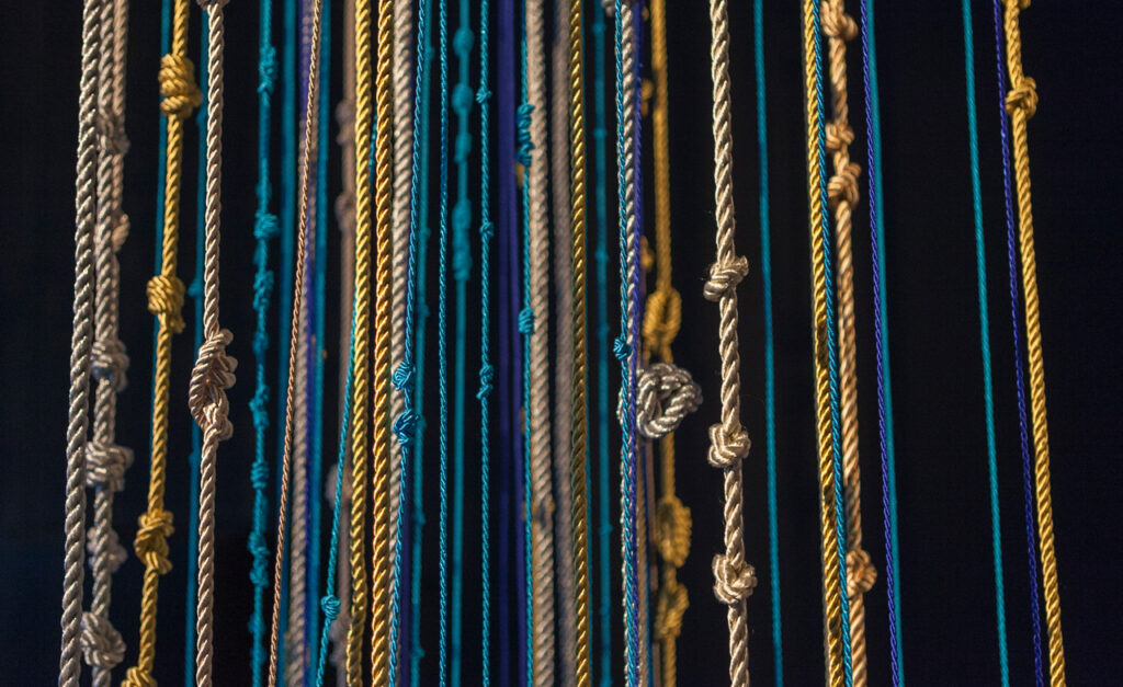 Tens of silken knotted ropes in shades of gold and blue hang vertically