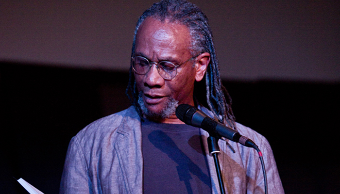 Nathaniel Mackey wearing a grey jacket and braided hair reads at a mic