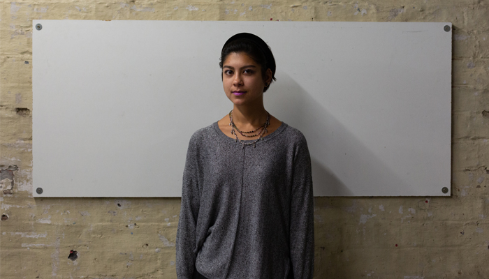 Nisha stands against a white wall, they are wearing a grey jumper