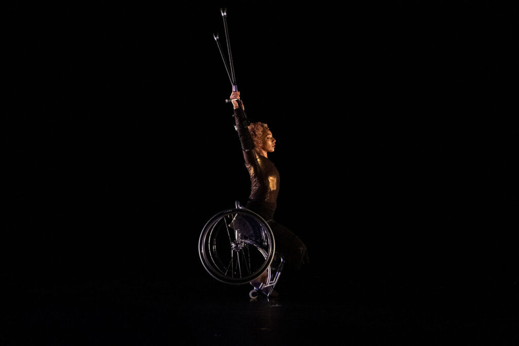 Alice is centre stage in her manual wheelchair, arms raised, crutches extended