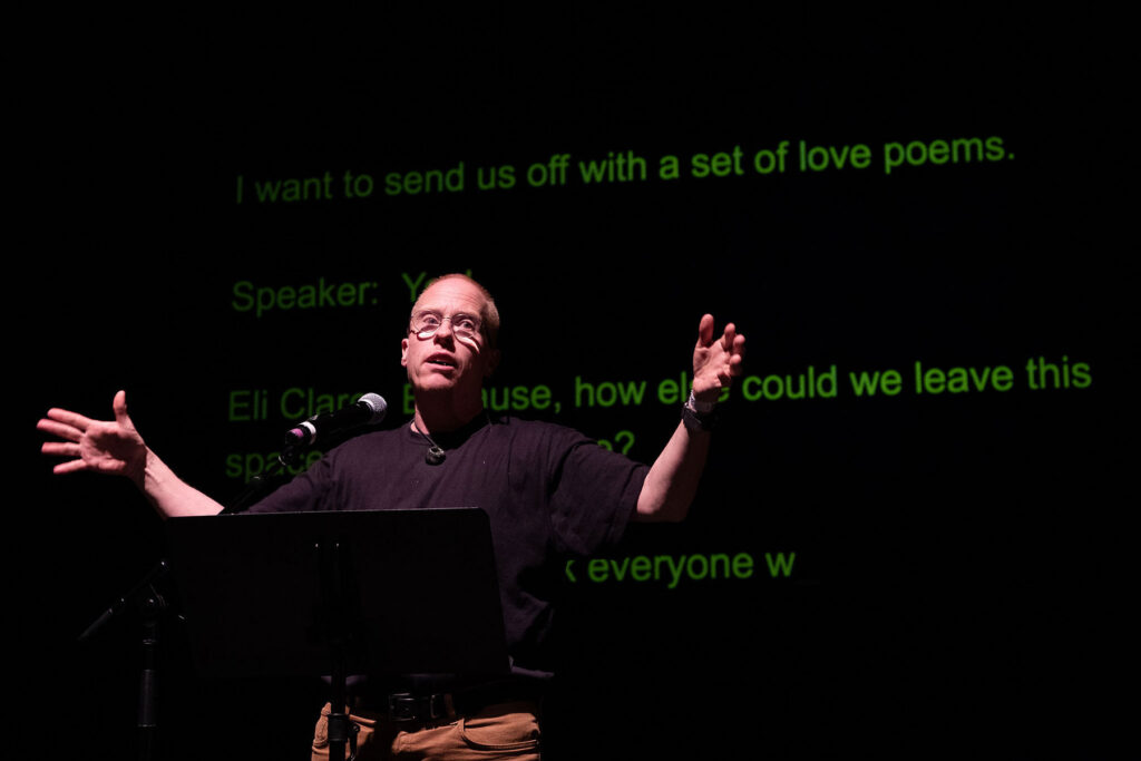 Eli Clare standing on a stage, arms outstretched