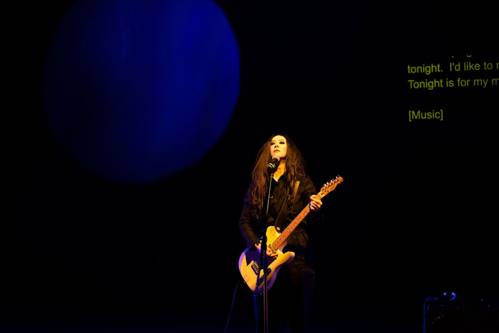 Johanna Hedva standing on a darkened stage, guitar in hand.