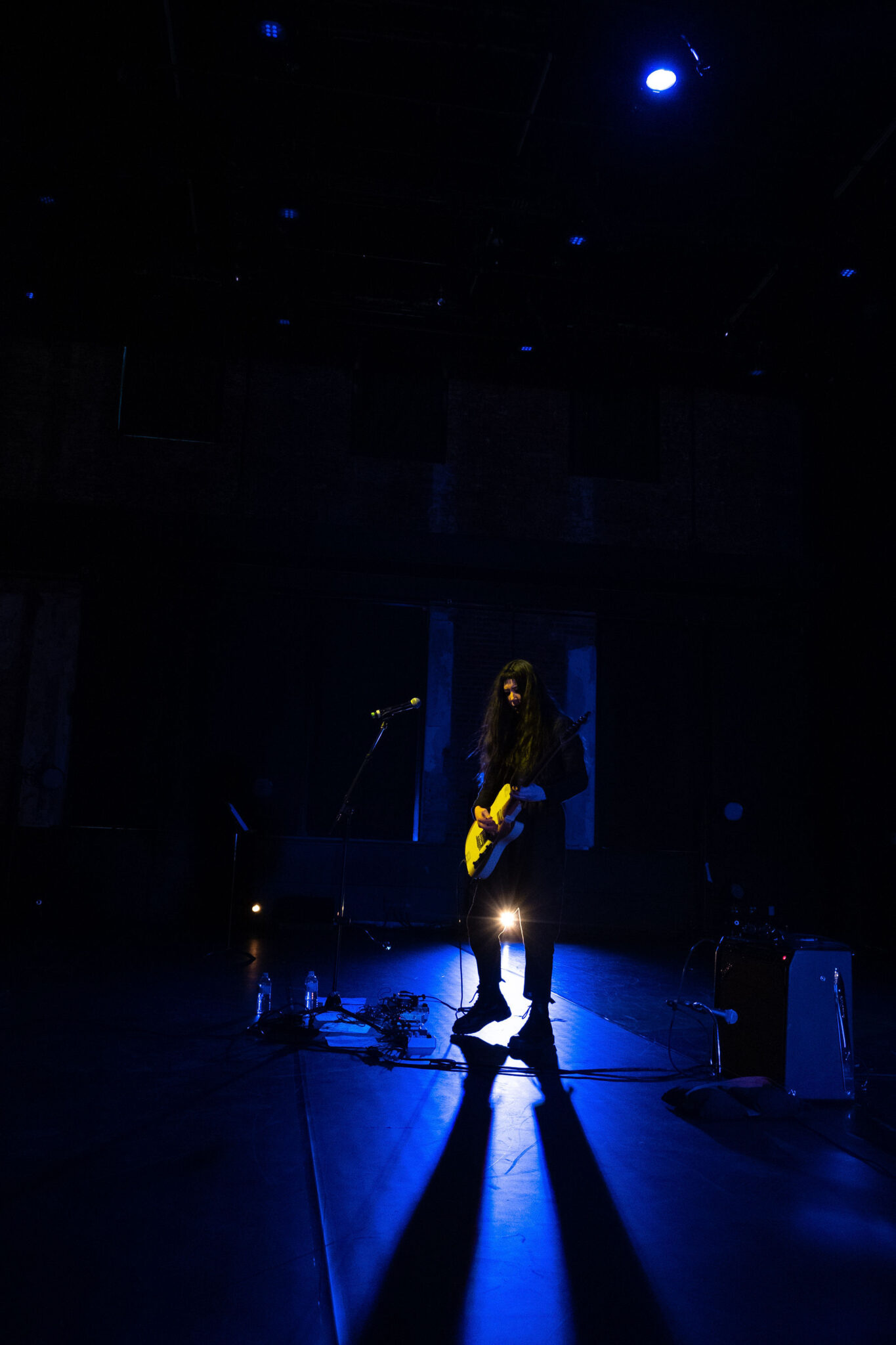 Johanna Hedva standing on a dark stage with a guitar, backlit in blue light, 