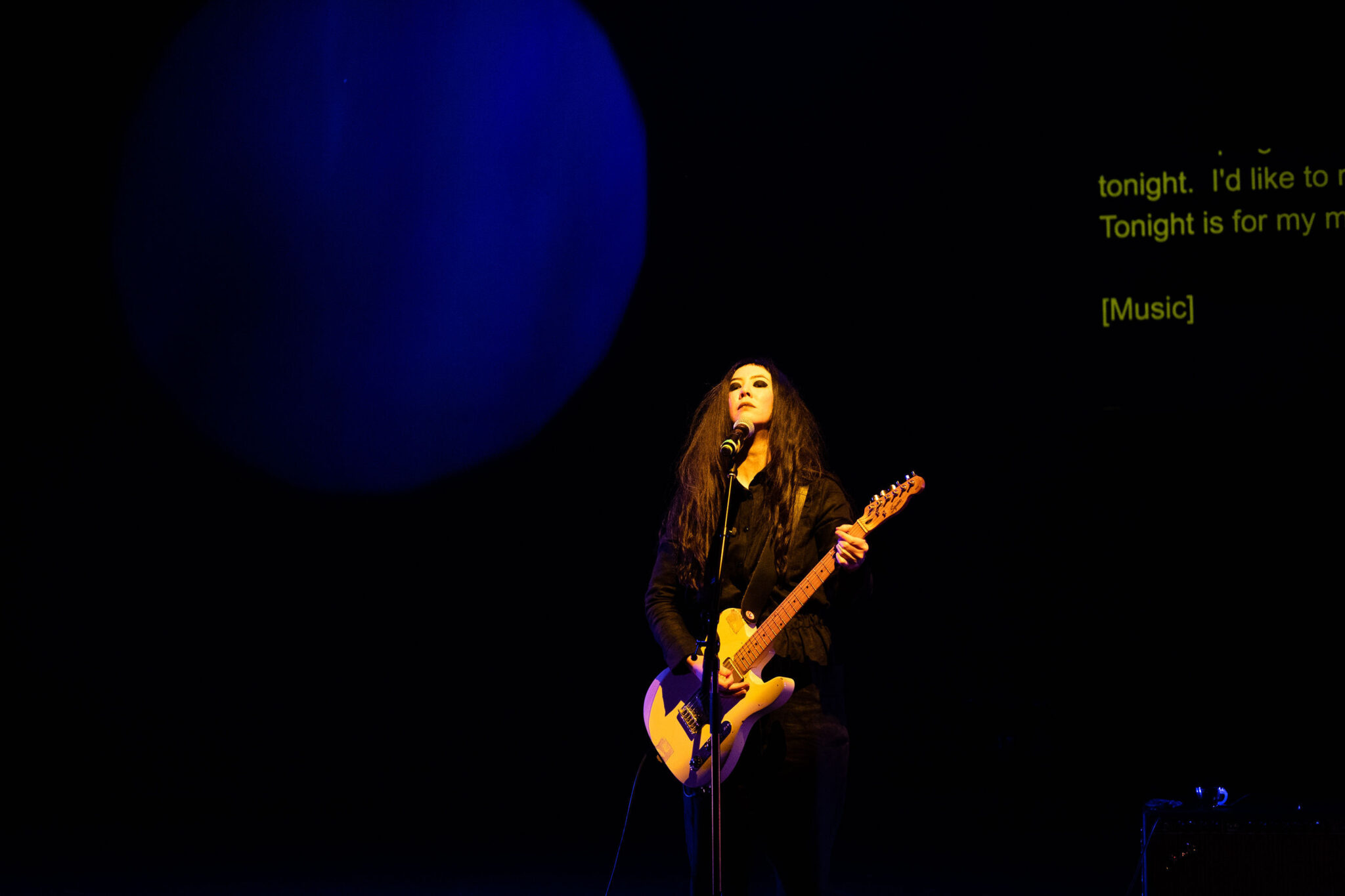 Johanna on a dark stage, dimly lit with a yellow spotlight, guitar in hand