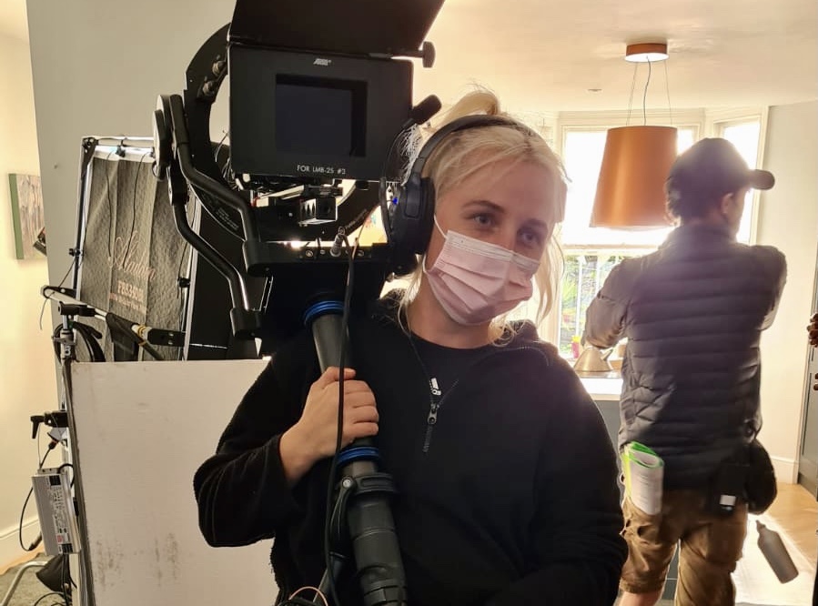 Lou, a white woman in her 20s with bleached blond hair, carries a huge camera on a film set, she is wearing a pink face covering and a black hoody