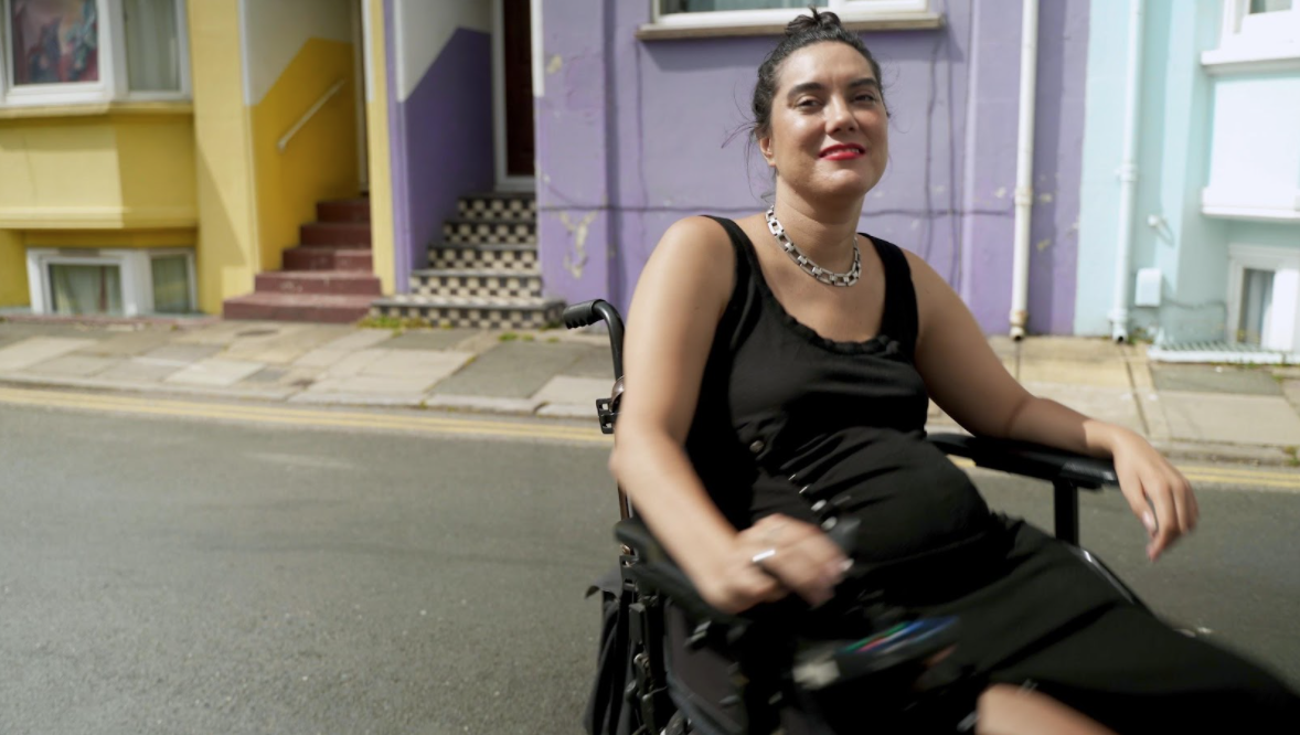 In front of bright pastel coloured houses, a sun kissed Kyla, a disabled, mixed heritage woman in her 30’s, spins in her power chair and she wears red lipstick, a thick chain necklace and a black dress. Although dynamic and mid-spin, she is relaxed but gleeful.
