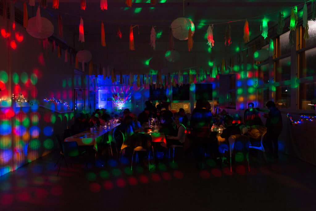 people sitting at tables within a dark room, lit with brightly coloured fairy lights