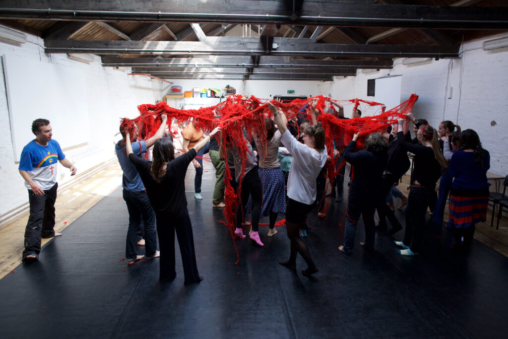 a room full of people standing in a big huddle, everyone is holding a massive, tangled bunch of red wool which seems to be connecting and entangling everyone.