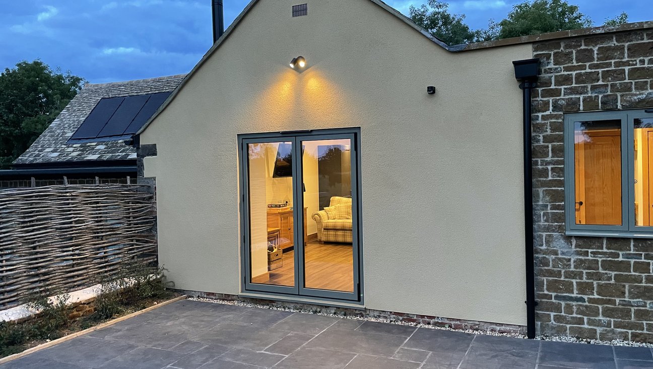 

                                        a rendered gable end with double doors on the left, a stone square extension with a window in to the right. in the dusk looking in to the cosy living area through the window.