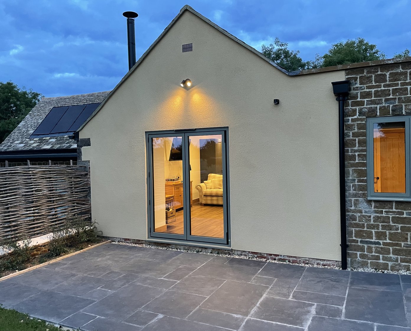a rendered gable end with double doors on the left, a stone square extension with a window in to the right. in the dusk looking in to the cosy living area through the window.