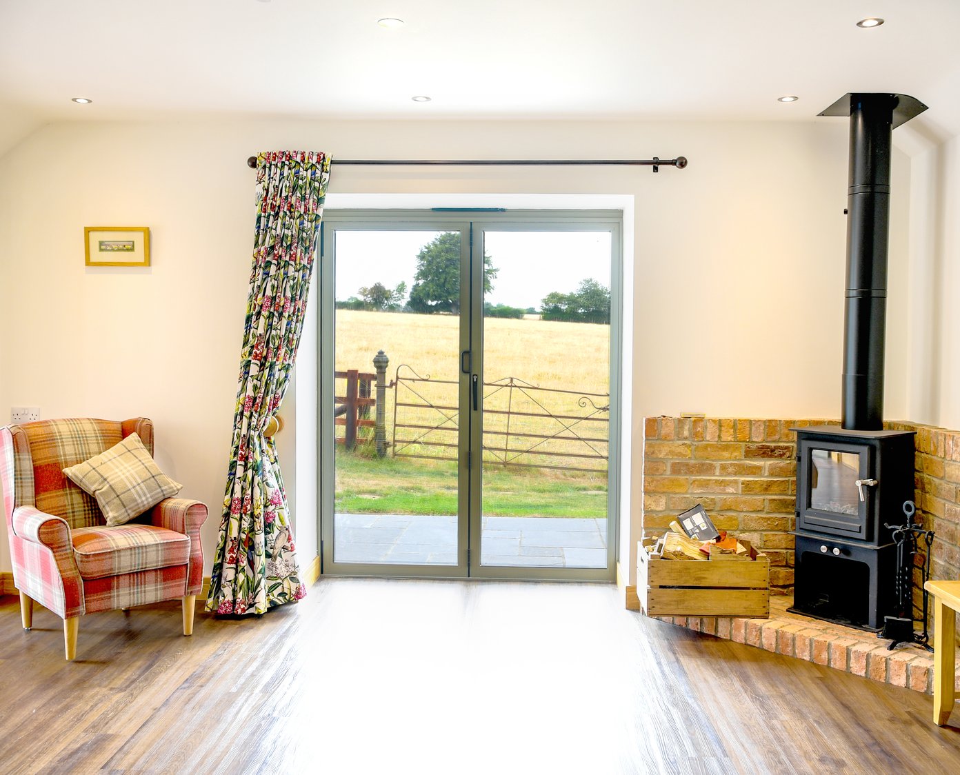 Tartan Chair on the left of a patio window, with a brick fireplace and logburner to the right. the view out of the window is out on to parkland with old wrought iron gate in the foreground