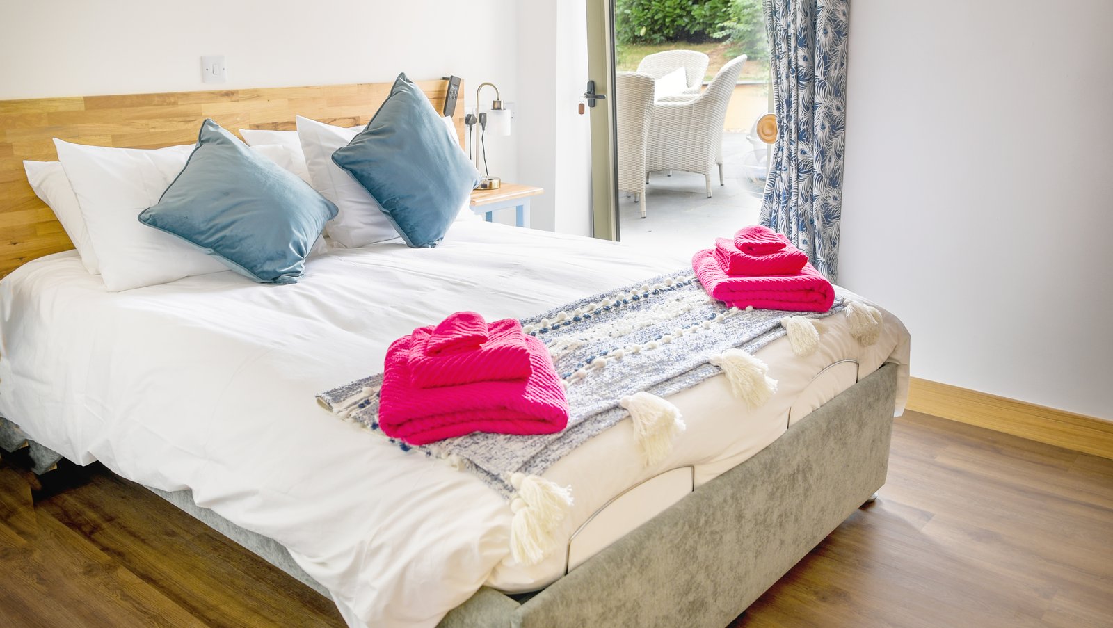 profiling bed kingsize bed in white room with blue throw and cushions; Bright pink towels on bed. green floor to ceiling window with feather pattern blue and cream curtain.