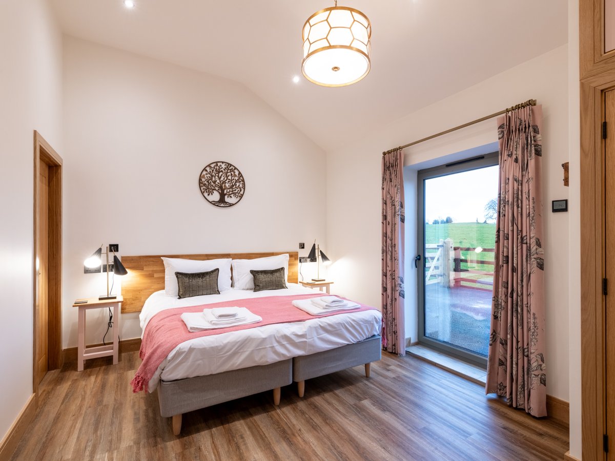 A generous bedroom in white with a non slip oak effect floor. Super king bed made up in white cotton linen with pillows, green cushions and a pink blanket. a floor to ceiling window and built in oak wardrobe to the left.