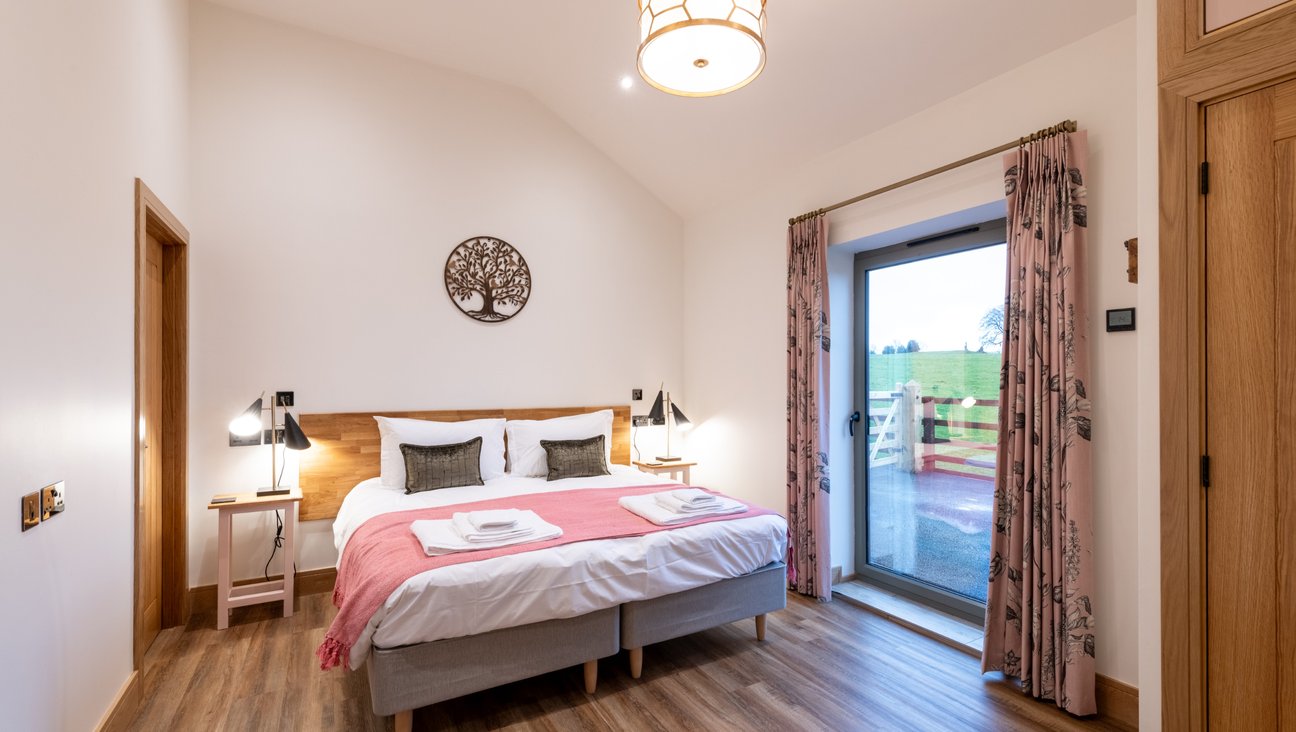 

                                        A generous bedroom in white with a non slip oak effect floor. Super king bed made up in white cotton linen with pillows, green cushions and a pink blanket. a floor to ceiling window and built in oak wardrobe to the left.