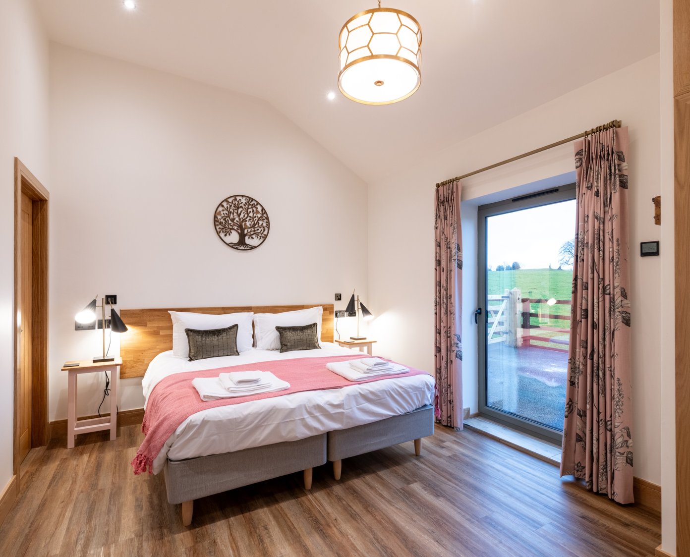 A generous bedroom in white with a non slip oak effect floor. Super king bed made up in white cotton linen with pillows, green cushions and a pink blanket. a floor to ceiling window and built in oak wardrobe to the left.