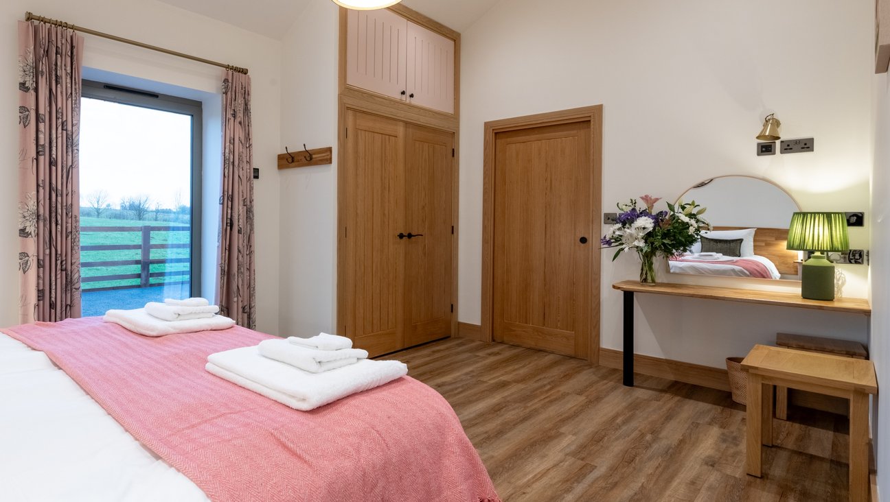 

                                        Super King bed to the left in front of window looking out to field, built in wardrobe, sliding door and dressing table with green lamp and flowers. The room has ample space for a wheelchair