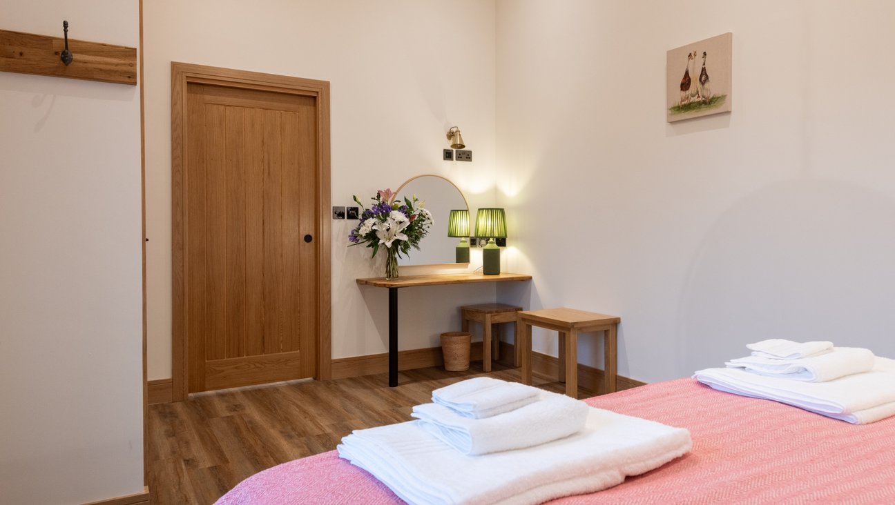 

                                        Large Bedroom with sliding oak door, built in oak dressing table and a side table to hold other bits and bobs. to the left is a built in wardrobe with robe hooks and in the foreground a super king bed with fluffy white towels on it