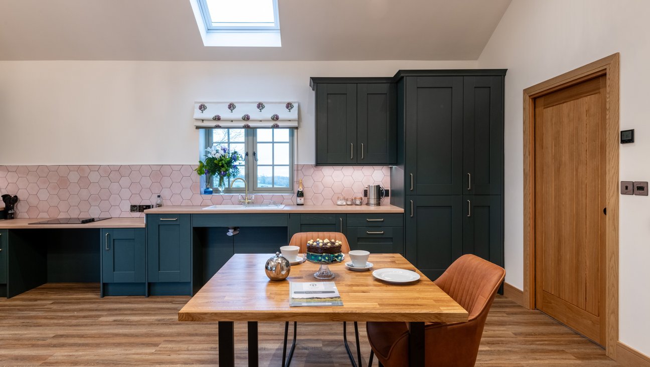 

                                        green floor to ceiling kitchen in back ground with wheel chair friendly roll under sink and hob. Oak dining table and leather chairs in foreground