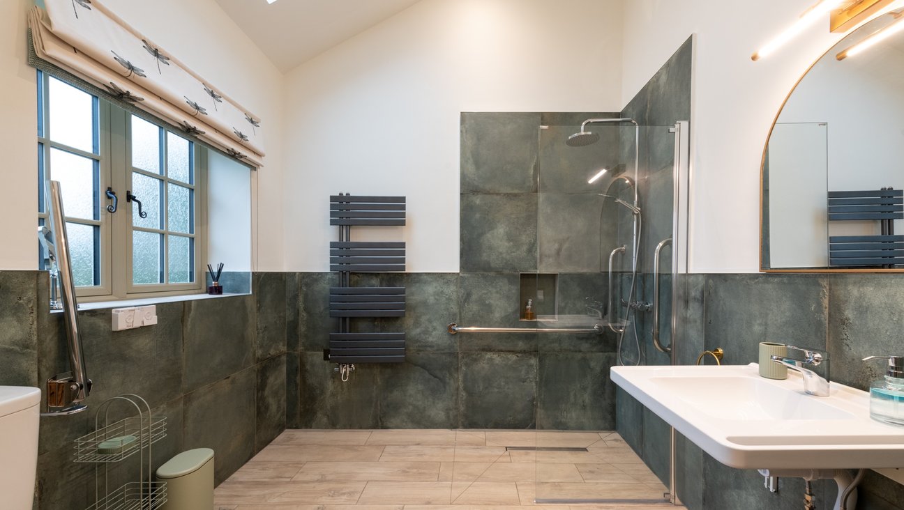 

                                        accessible shower room tiled in green, with a non slip oak effect tiled floor. hewi sink with integrated grab bars and gold framed mirror. E shaped heated towel rail. comfort height toilet with two drop down grab rails.