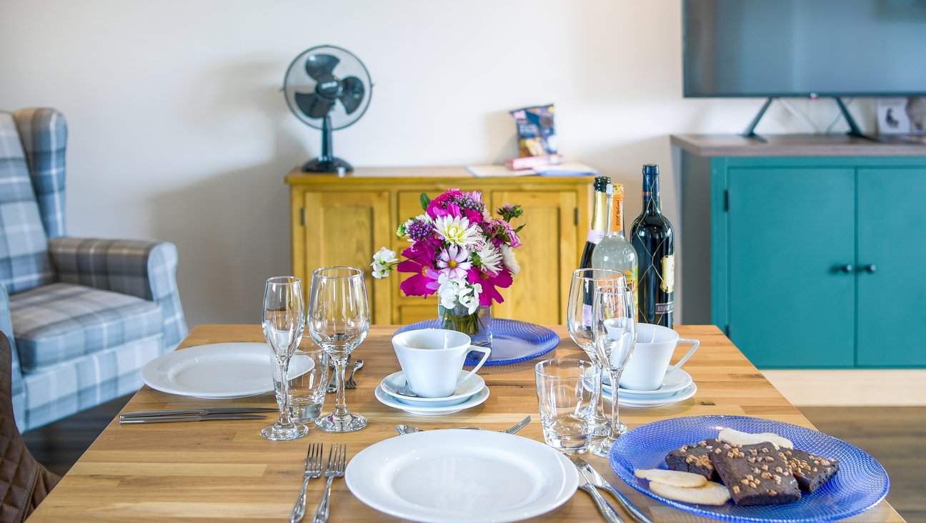 

                                        Oak table set with pink flowers cutlery, cakes and bottles. dark wood floor in a sunny white room with a blue kitchen to the left of the image