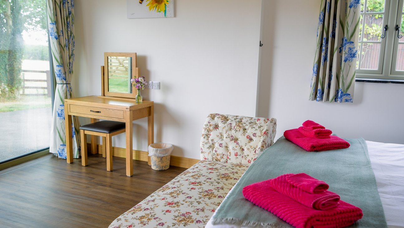 

                                        Bed to right hand side in white room with wood floor and bright pink towels on it. on the left window looking out on to parkland and an oak dressing table and seat