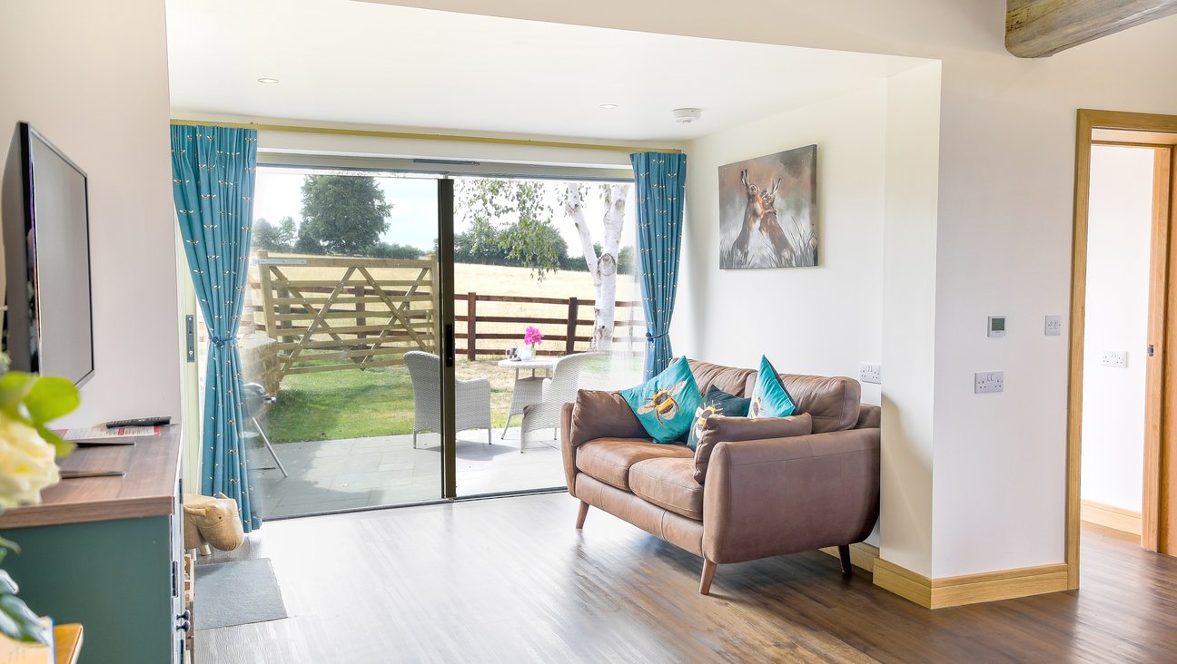 

                                        Living area with large windows on to a view of parkland. a leather sofa on the right hand side of a white room and a logburner on a slate fireplace on a dark wood floor. it is a sunny day