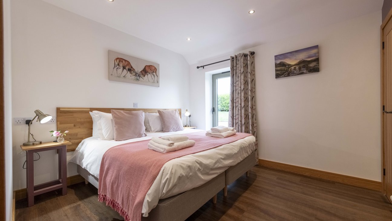 

                                        twin beds with oak headboard, made up with pure white cotton linnen, and dusky pink throws and cushions. bright pink towels on the bed, in background large green framed floor to ceiling window with feather patterned curtain.