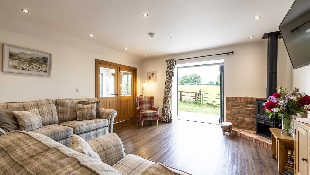 

                                        A view of fields out of bifold doors, with a tweed armchair to the left and log burner to the right. the floor is level and made of warmwood, with level access to a slate patio beyond.