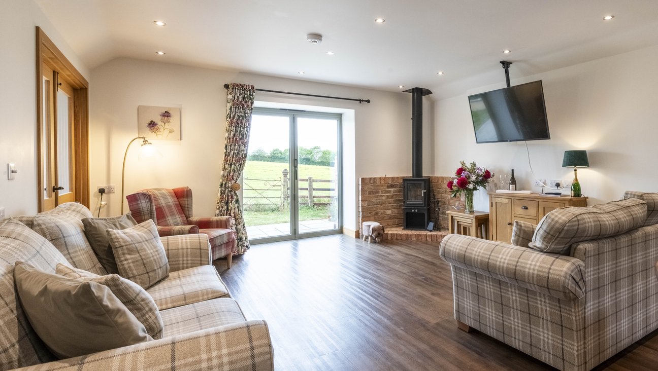 

                                        Tartan Chair on the left of a patio window, with a brick fireplace and logburner to the right. the view out of the window is out on to parkland with old wrought iron gate in the foreground