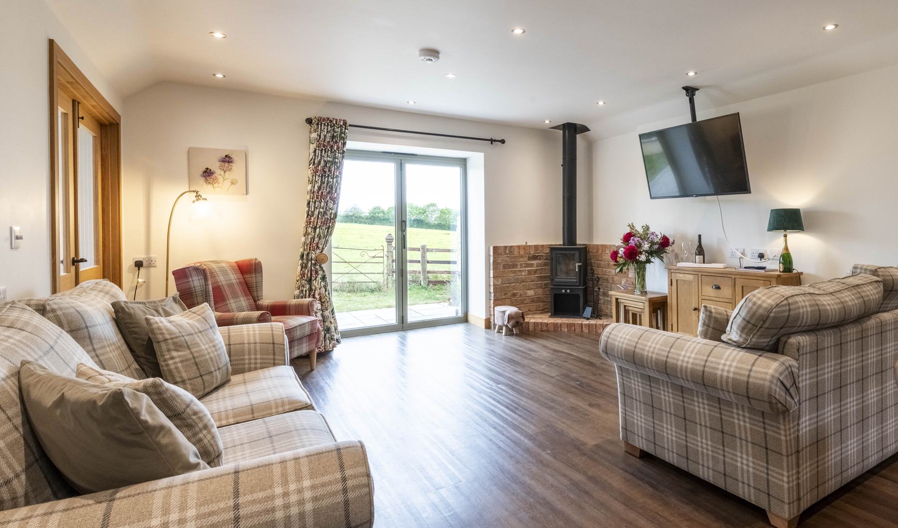 Tartan Chair on the left of a patio window, with a brick fireplace and logburner to the right. the view out of the window is out on to parkland with old wrought iron gate in the foreground