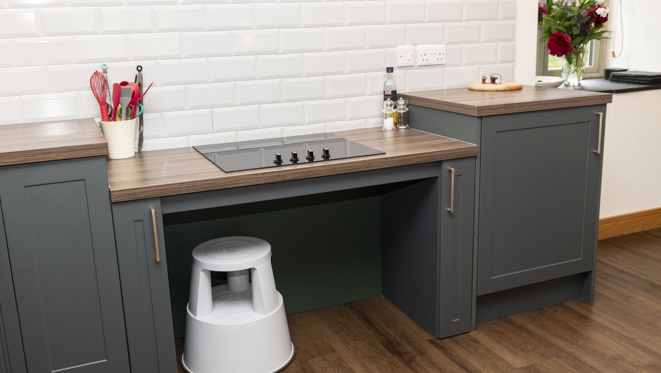 

                                        Black roll under ceramic hob with front controls, good grips utensils in a white jar to the left of hob, and toaster in back left hand corner, gray brick style tiles on the wall