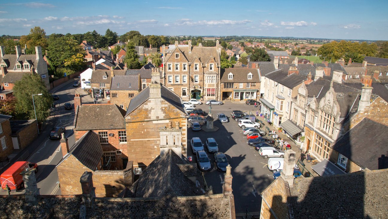 

                                        View from the air of a traditional market place. A beautiful coaching in built in stone the colour of gingerbread, pubs, cafes and shops surround parked cars in the centre.
