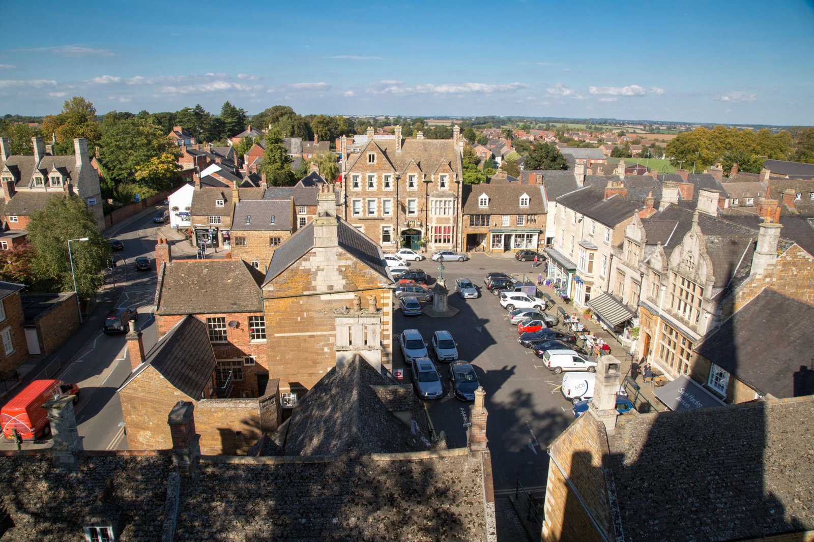 View from the air of a traditional market place. A beautiful coaching in built in stone the colour of gingerbread, pubs, cafes and shops surround parked cars in the centre.